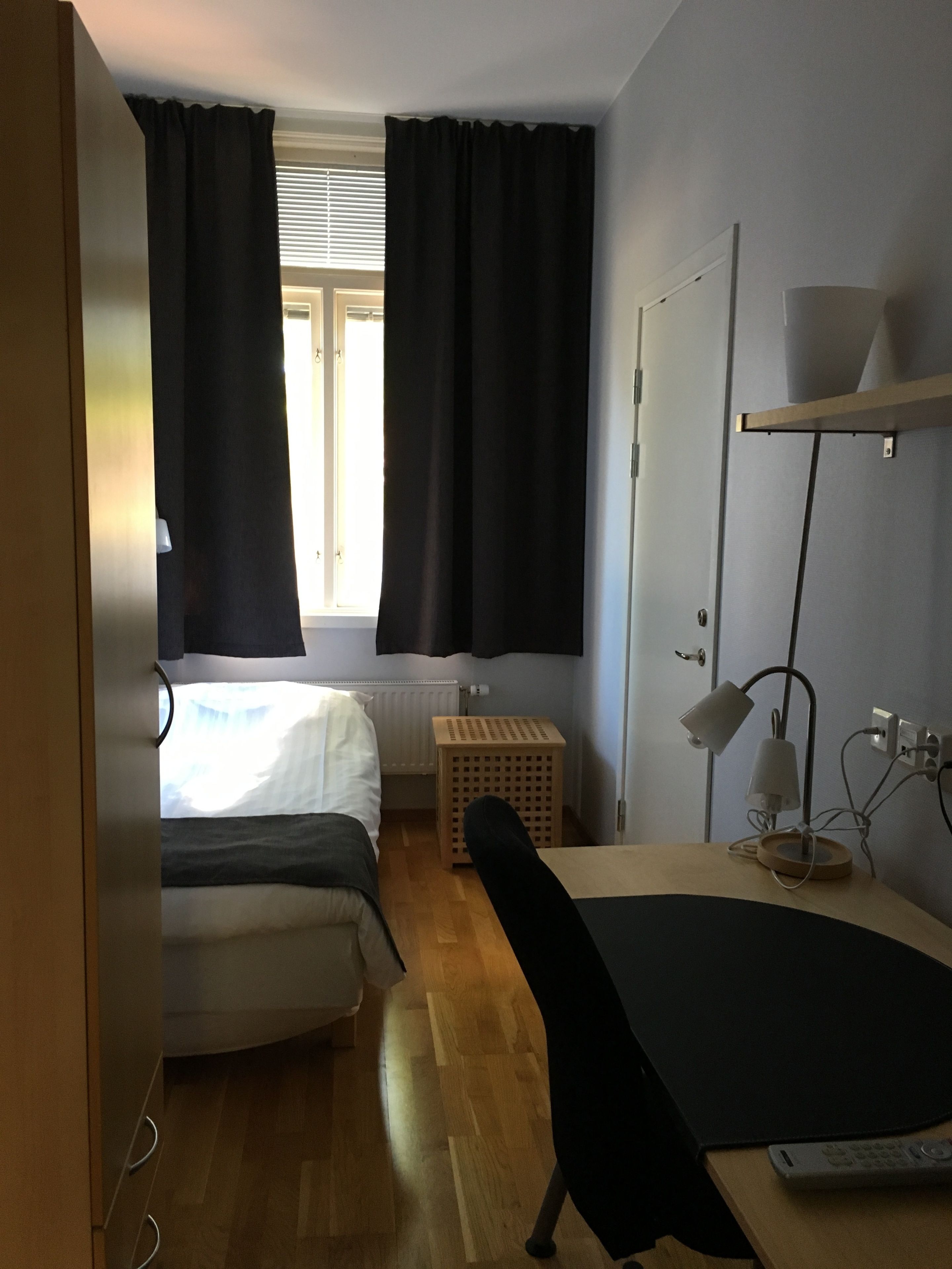 A room with two beds, a desk with a lamp, a radiator, a window with curtains, and a wooden floor.