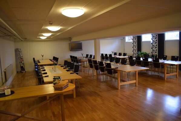 A conference room with multiple tables and chairs, a flat-screen TV, and a large window with blinds. - Hotell Hässlö (Vasteras)