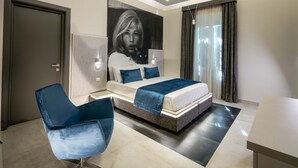A modern bedroom with a bed, a blue chair, a large painting, and a desk. - Via Veneto Luxury Suites (Rome)