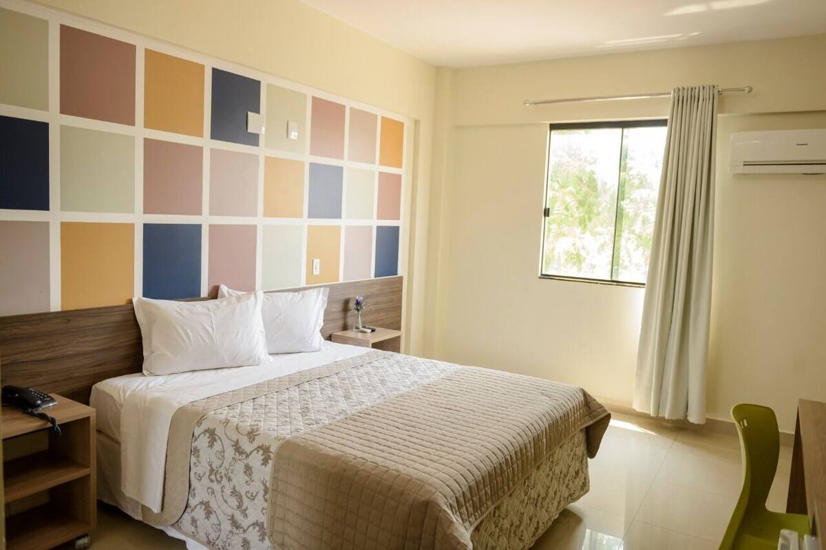 A hotel room with a large bed, a bedside table with a lamp, a window with curtains, and a wall with a colorful panel design.