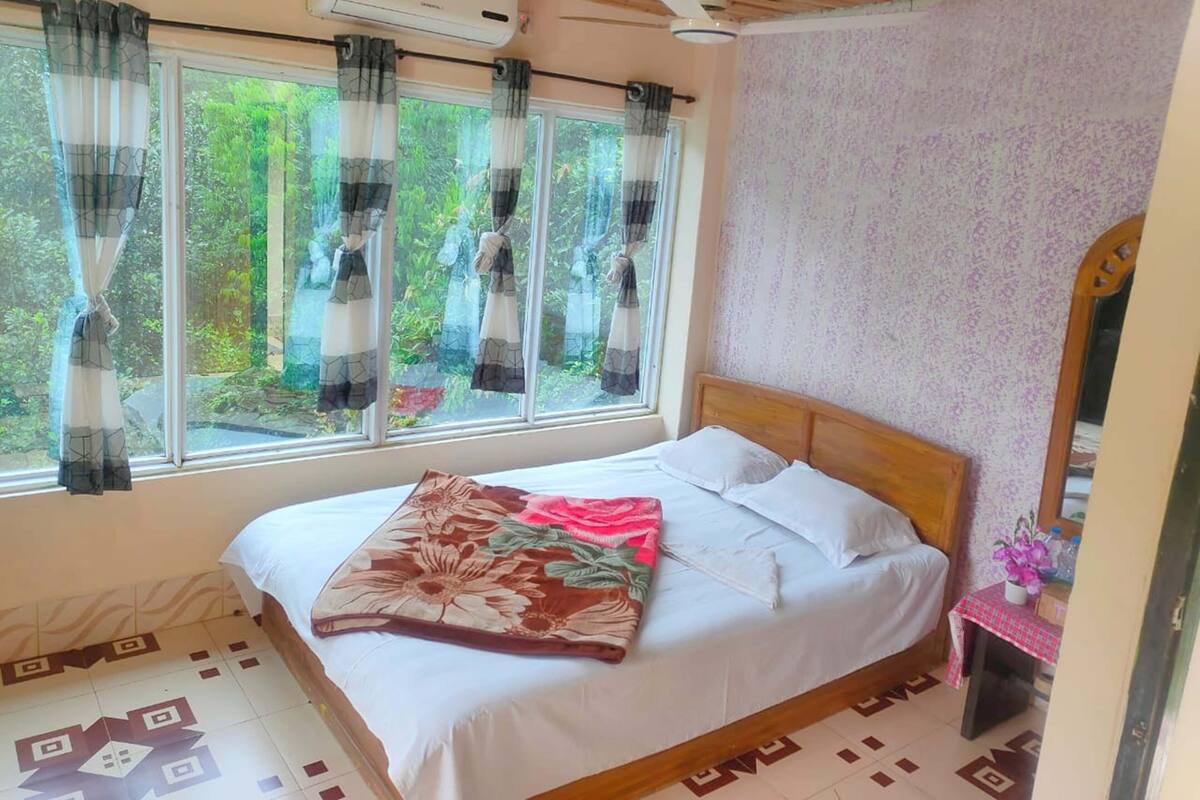 Un dormitori amb un llit, una finestra amb cortines, un aparell d'aire condicionat, un ventilador de sostre i una taula petita amb un mirall i flors.