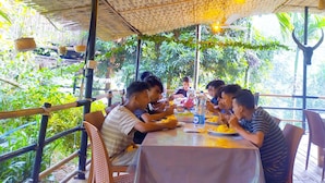 Een groep mensen dineert aan een buitentafel onder een rieten dak, omgeven door weelderige begroeiing.
