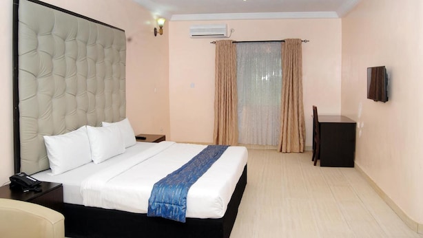 Quarto de hotel com uma cama grande, uma mesinha de cabeceira com telefone, uma escrivaninha com cadeira e uma TV fixada na parede.