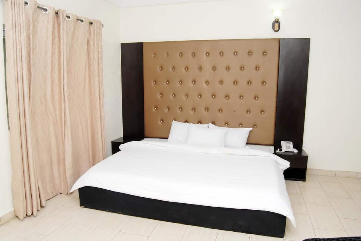 Ein Hotelzimmer mit einem großen Bett, einem gepolsterten Kopfteil, einem Nachttisch mit Telefon und einem Vorhang am Fenster.