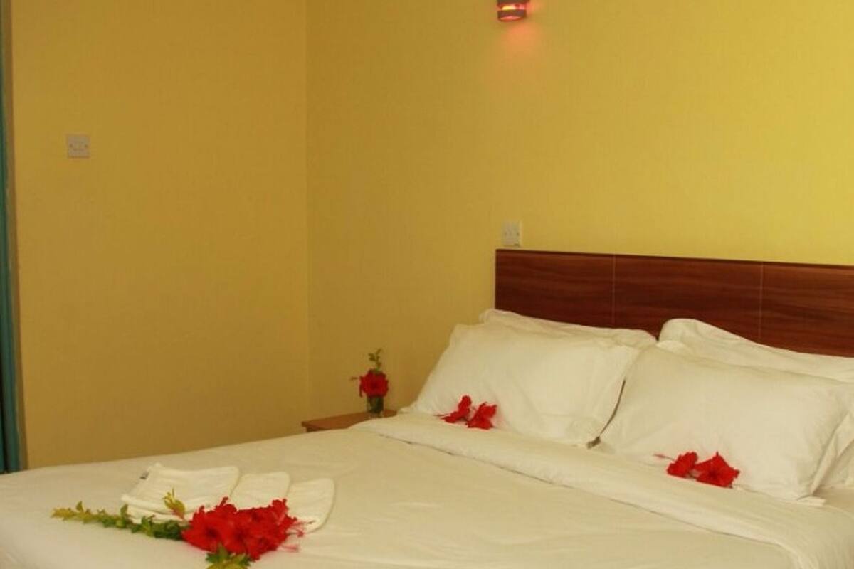 A neatly made bed with white linens and red flowers, a wooden headboard, and a small bedside table with a red flower vase.