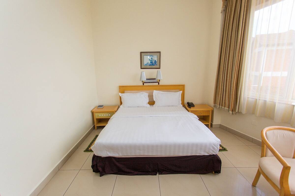 A hotel room with a bed, two bedside tables, a nightstand, a chair, and a window with curtains.
