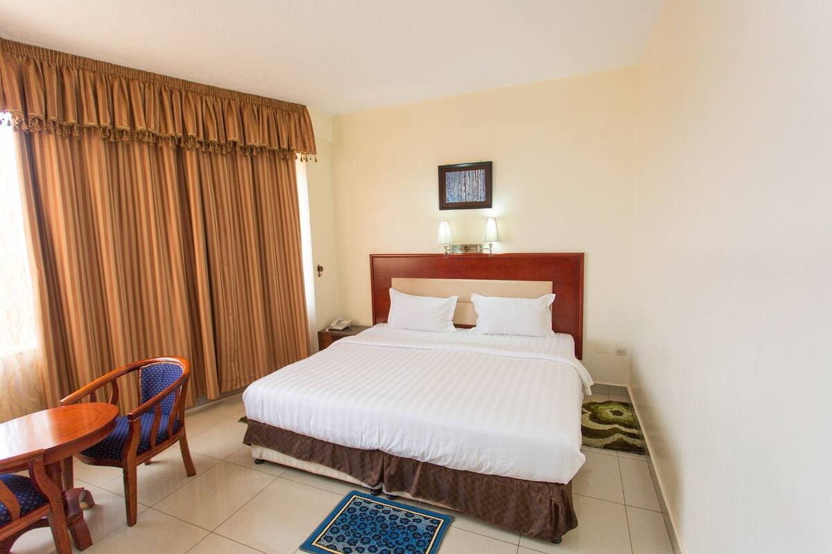 A hotel room with a large bed, a wooden chair, a nightstand, and a wall-mounted lamp.