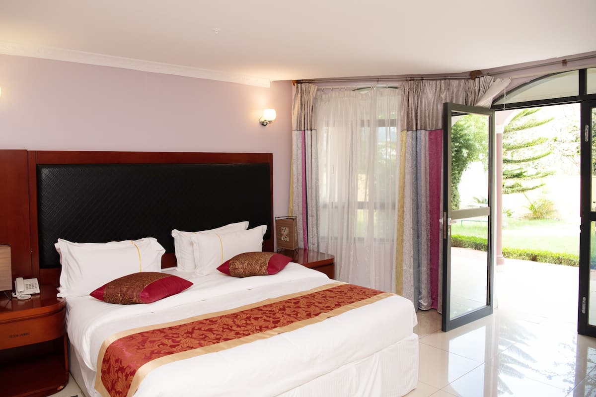 A hotel room with a large bed, a wooden headboard, a bedside table with a phone, and a view of a garden through a glass door.