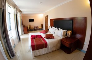 A hotel room with a large bed, a bedside table, a chair, a desk, and a TV mounted on the wall. - Orient Park Hotel (Kigali)