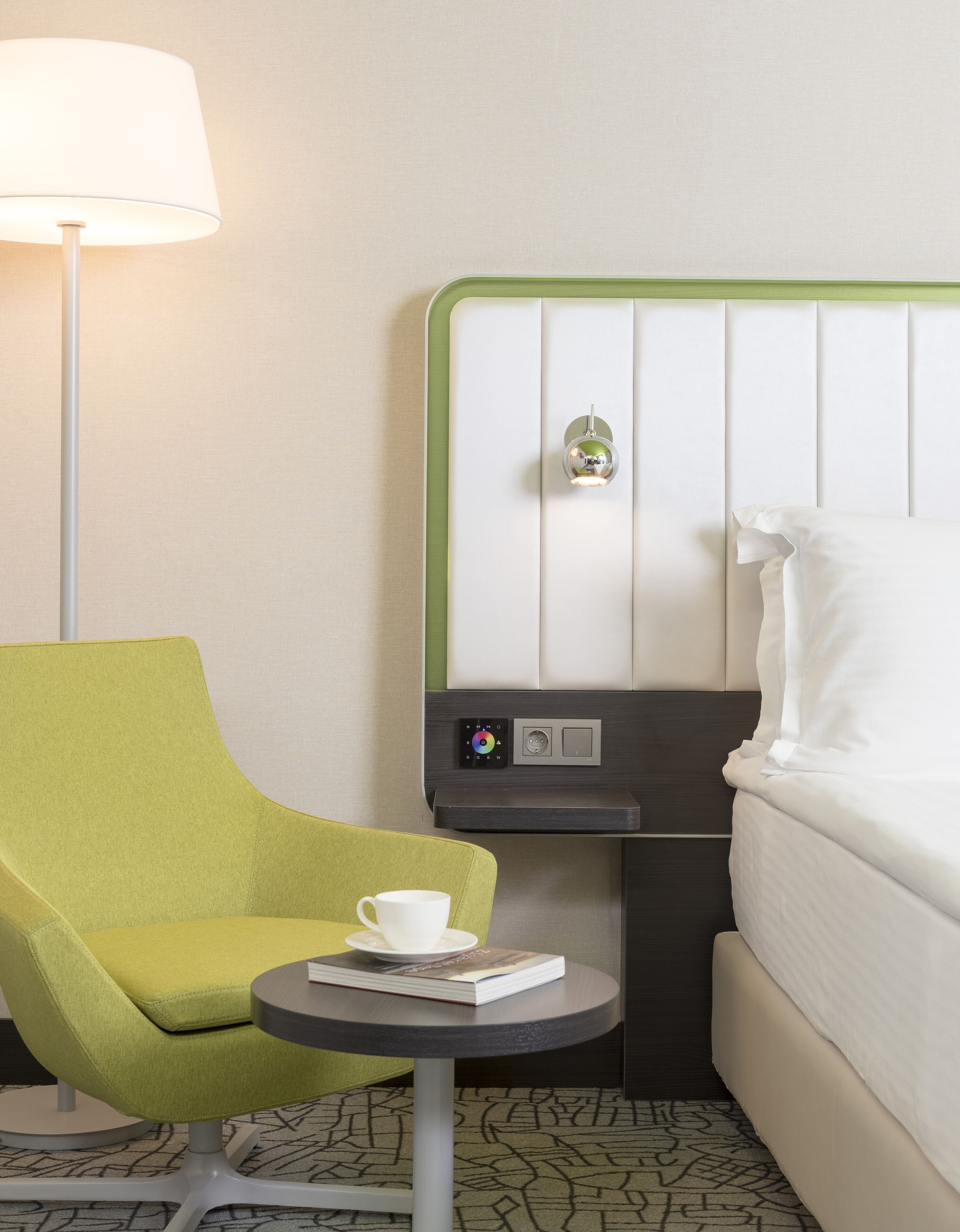 Habitación de hotel con un sillón verde, una mesita redonda con un libro y una taza, una cama con ropa blanca, un panel de control en la pared y una lámpara de pie.