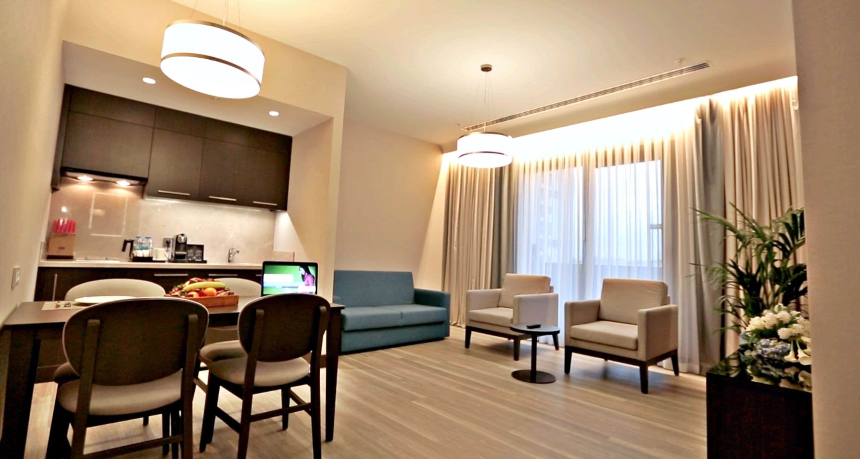 A modern hotel room with a dining area, a sofa, two armchairs, a small table, and a large window with curtains.
