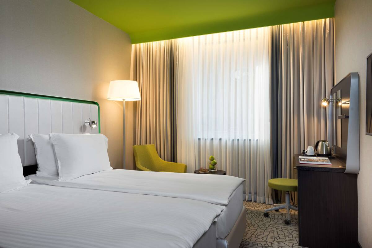 Et hotelværelse med en stor seng, et skrivebord med stol, en lampe og udsigt ud gennem gardiner.