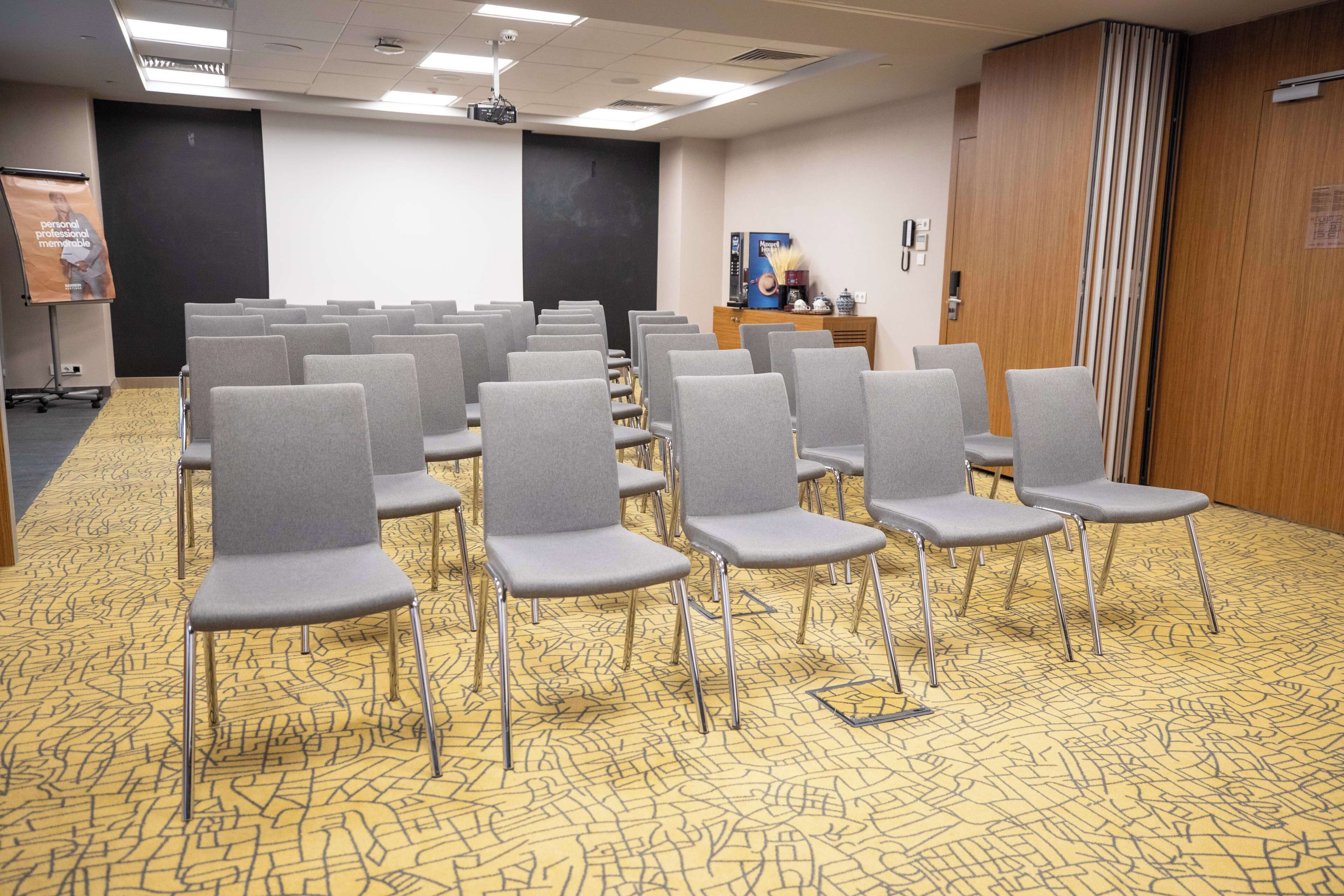A conference room with rows of grey chairs, a projector screen, a podium, and a carpeted floor.