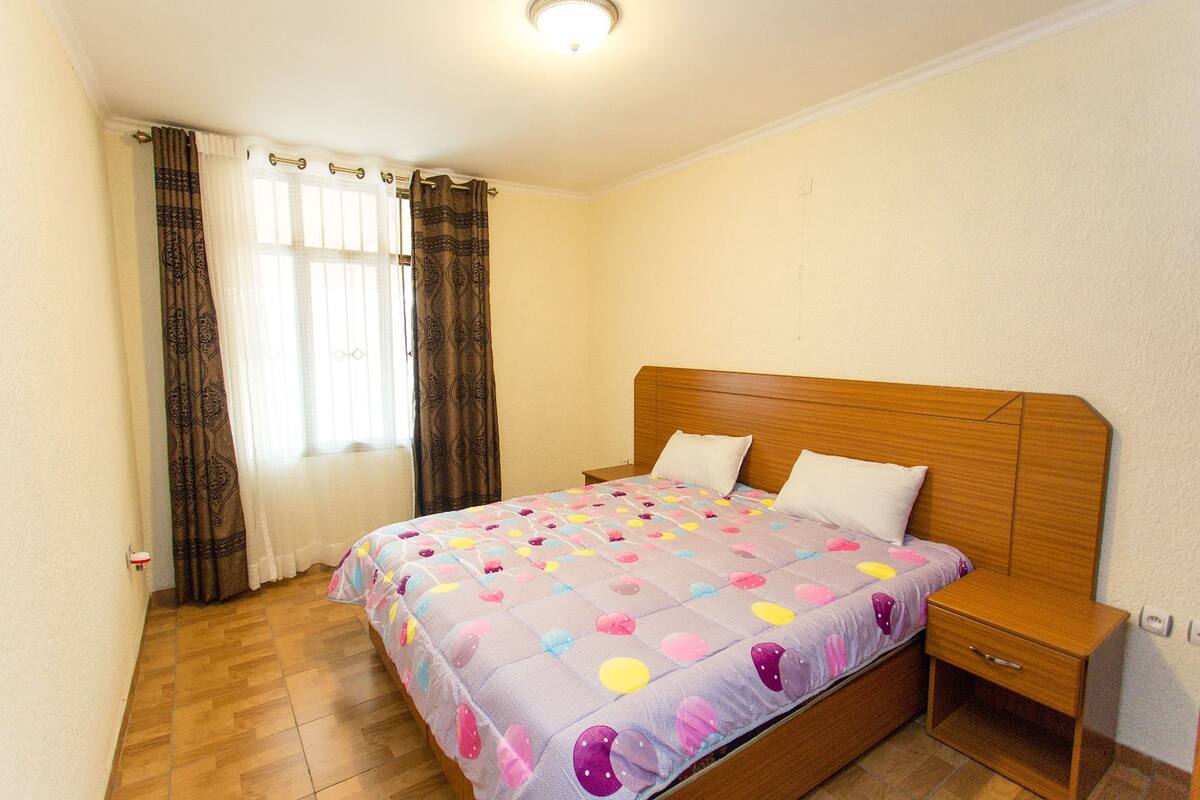 A bedroom with a wooden headboard, a bed with a colorful quilt, a bedside table, and a nightstand with a drawer.