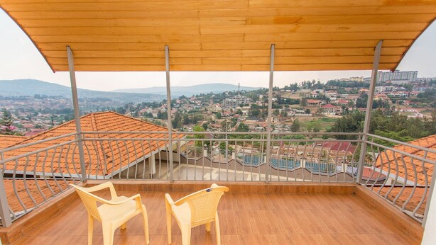 A balcony with two plastic chairs, a wooden ceiling, and a view of a town with red-roofed buildings. - Eden Escape Apartments (Kigali)