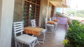 An outdoor patio with white chairs and tables, a metal grill, and a view of a hillside.