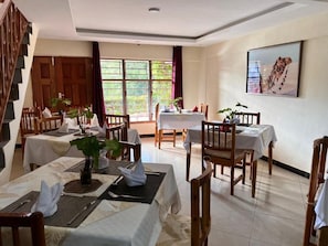 A dining area with tables set for guests, wooden chairs, and potted plants.