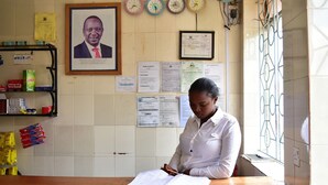 A person seated at a wooden desk with an open book, in a room with a tiled wall, clocks, framed certificates, and various notices. - Maru B Courts Hotel (Nyeri)