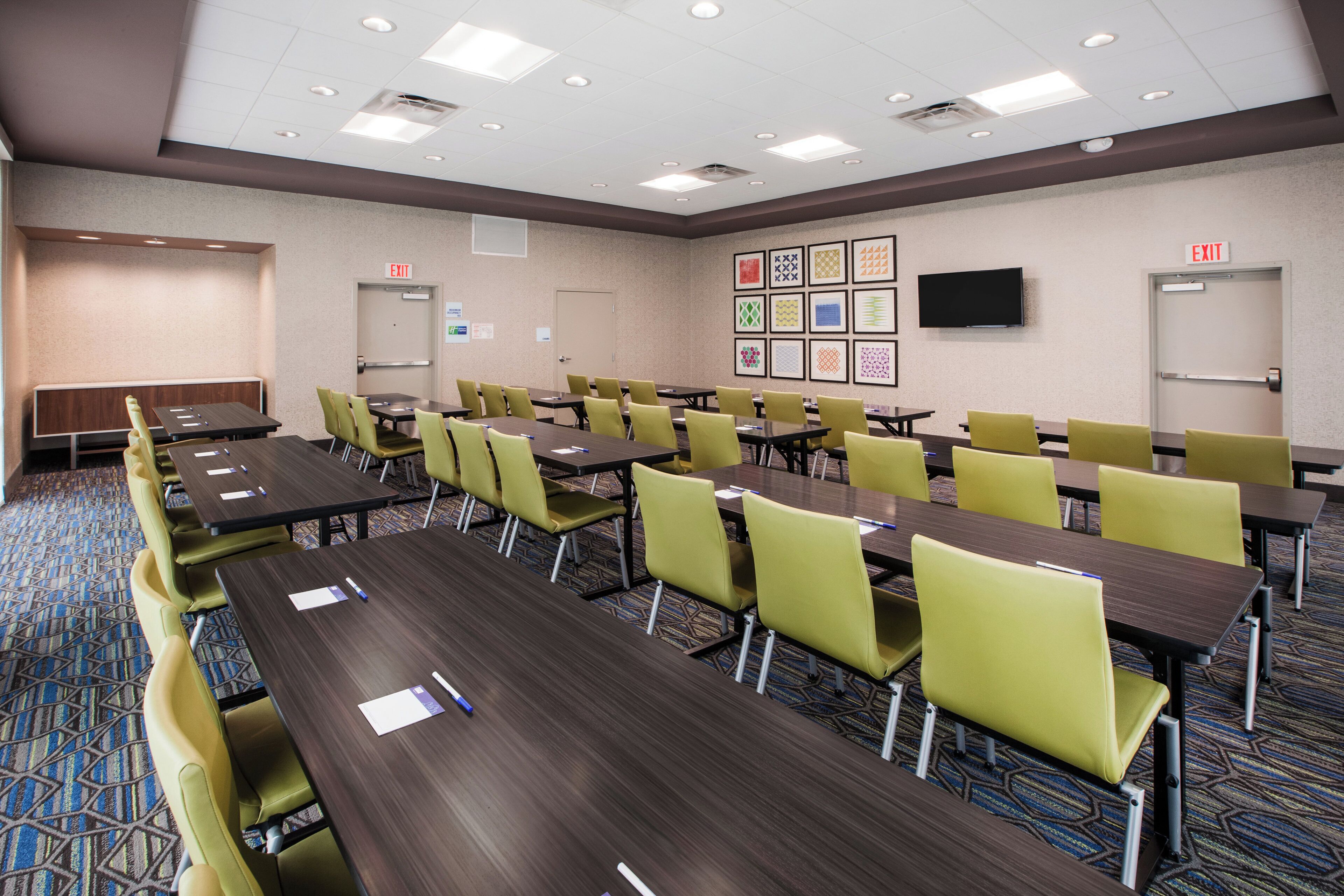 Une salle de conférence équipée de plusieurs tables et chaises, d’un téléviseur à écran plat fixé au mur et d’un panneau indiquant la sortie au-dessus de la porte.