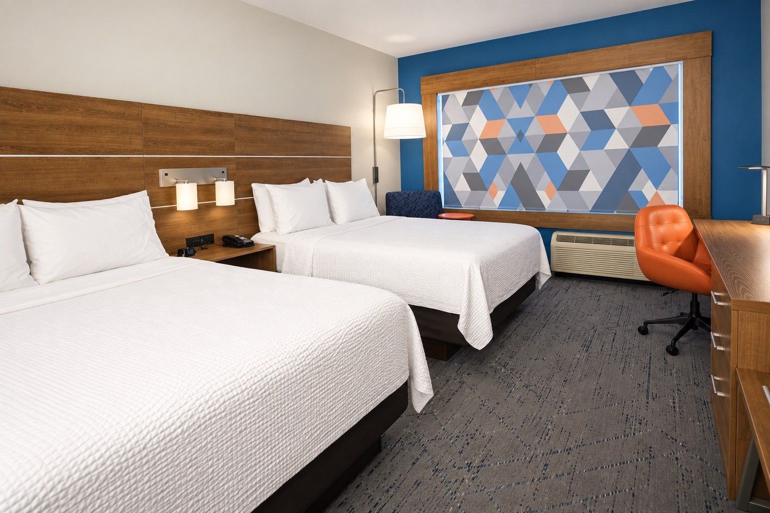 A hotel room with two beds, a desk, a chair, a large geometric wall art, and a lamp.
