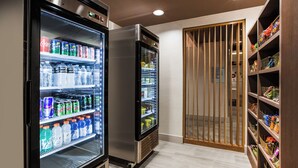 Ein Flur mit zwei Getränkeautomaten, gefüllt mit verschiedenen Getränken, einer Holztür mit Lamellen und einem Regal mit Snacks und anderen Artikeln.