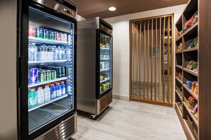Ein Flur mit zwei Getränkeautomaten, gefüllt mit verschiedenen Getränken, einer Holztür mit Lamellen und einem Regal mit Snacks und anderen Artikeln.