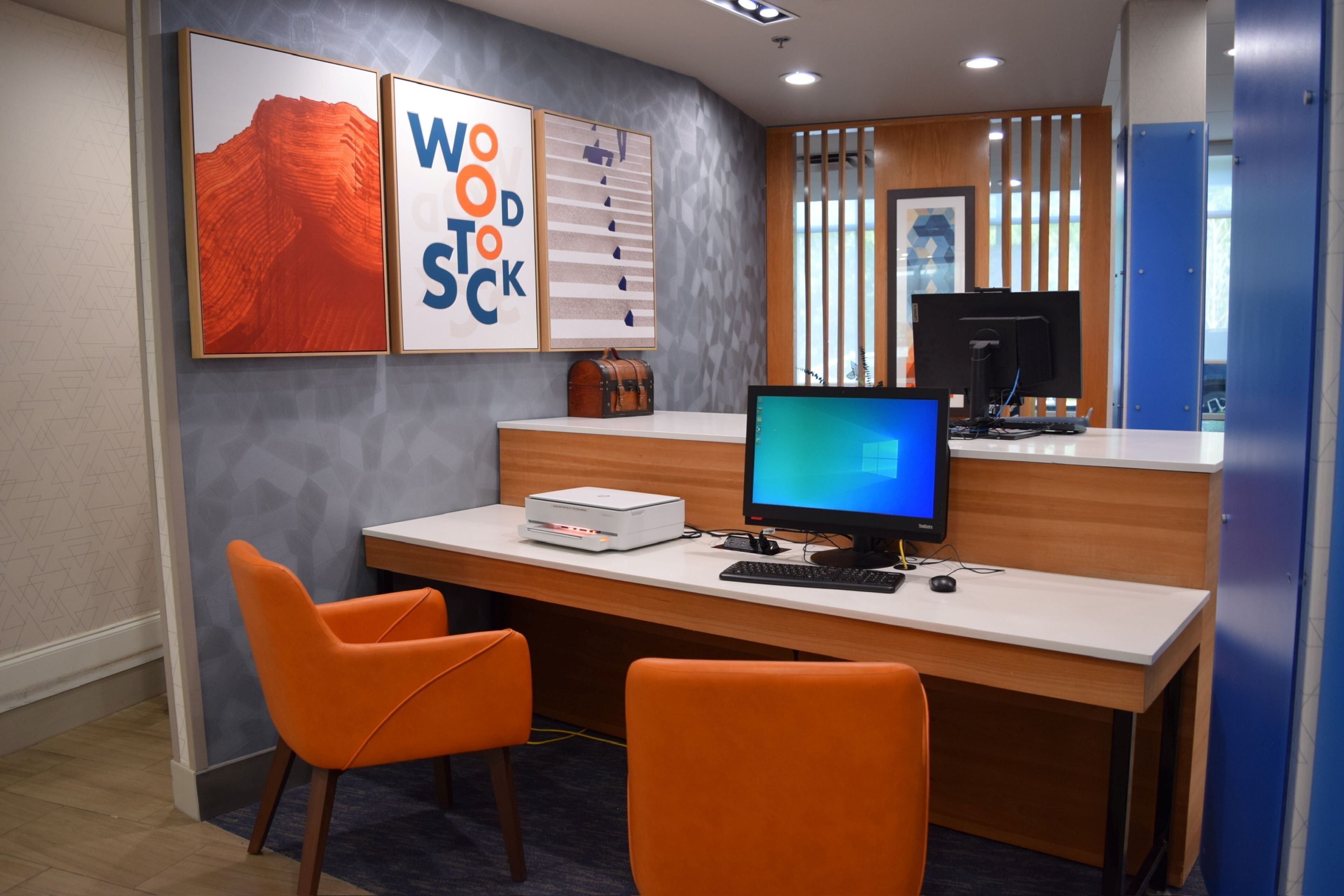 A modern office space with a desk, two orange chairs, a computer, and framed artwork on the wall.
