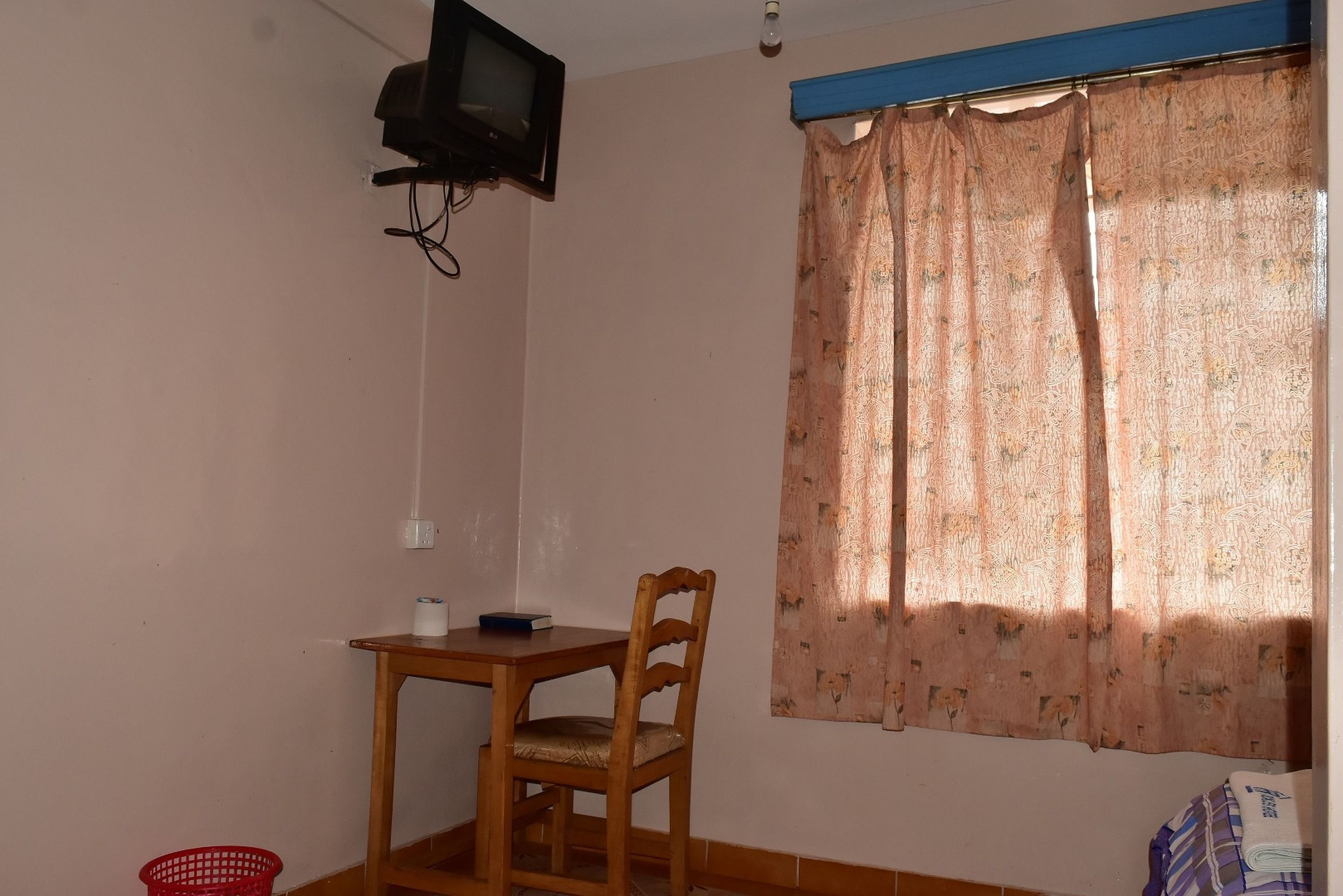 A small room with a wooden chair, a table, a television mounted on the wall, and a window with patterned curtains.