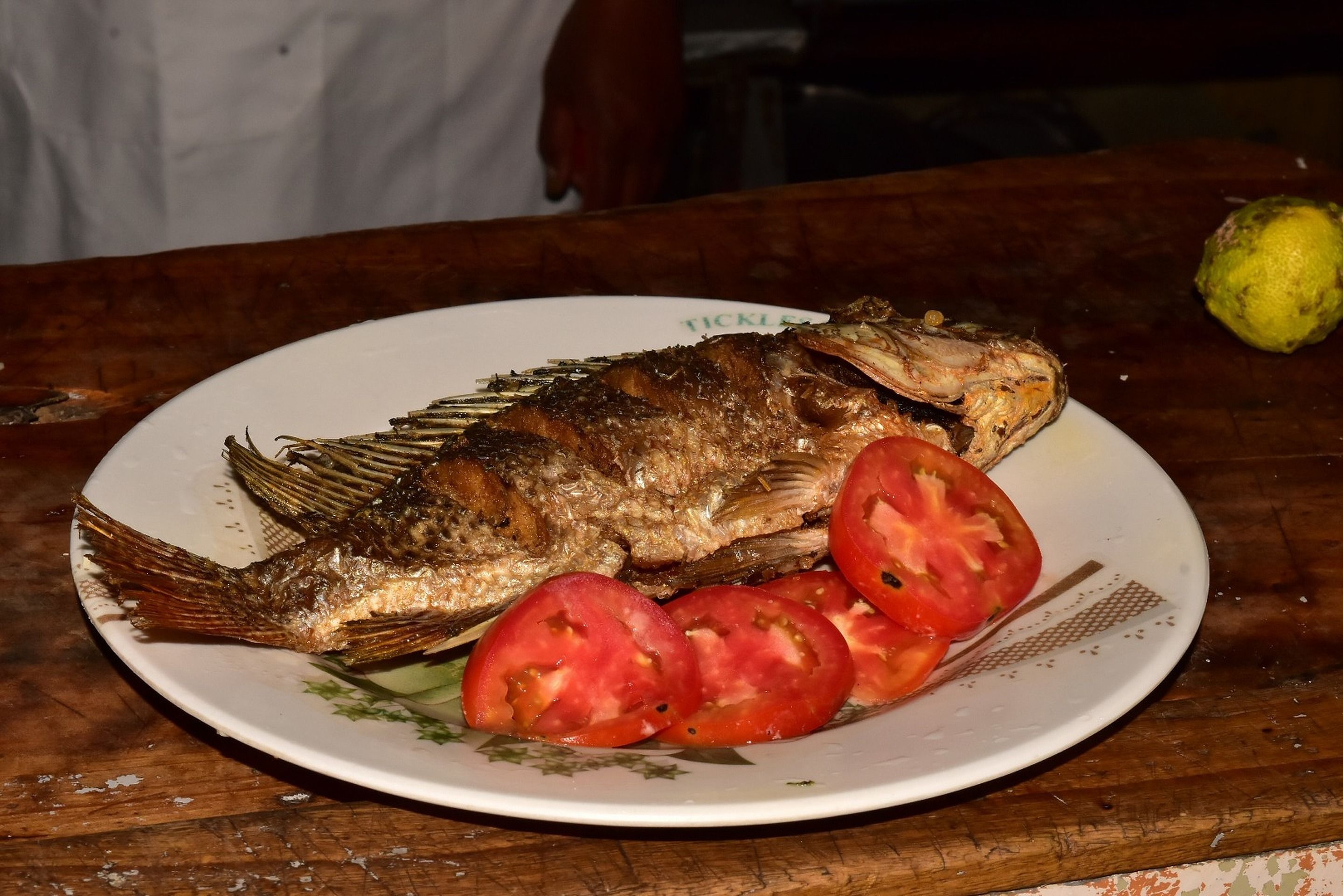 A plate with a whole fish, sliced tomatoes, and a lemon on a wooden table.