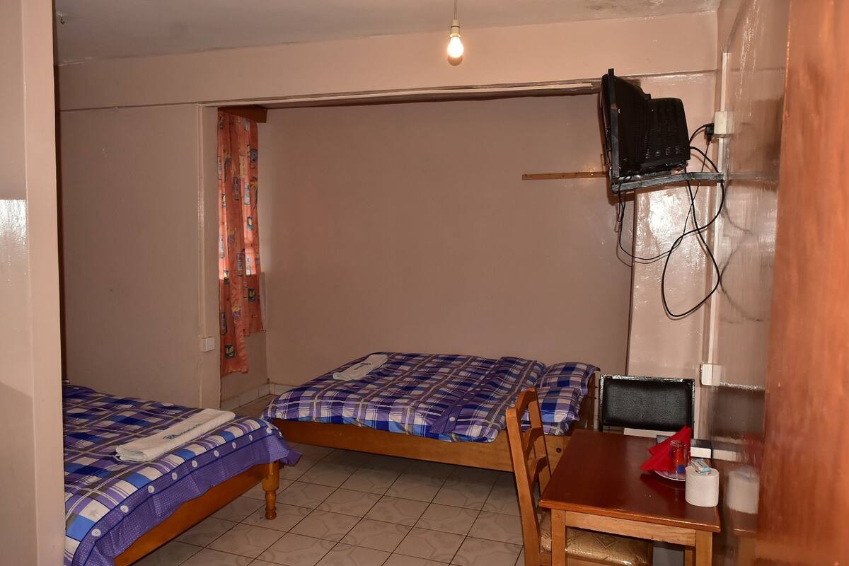 A small, compact room with two single beds, a wooden table, and a chair.