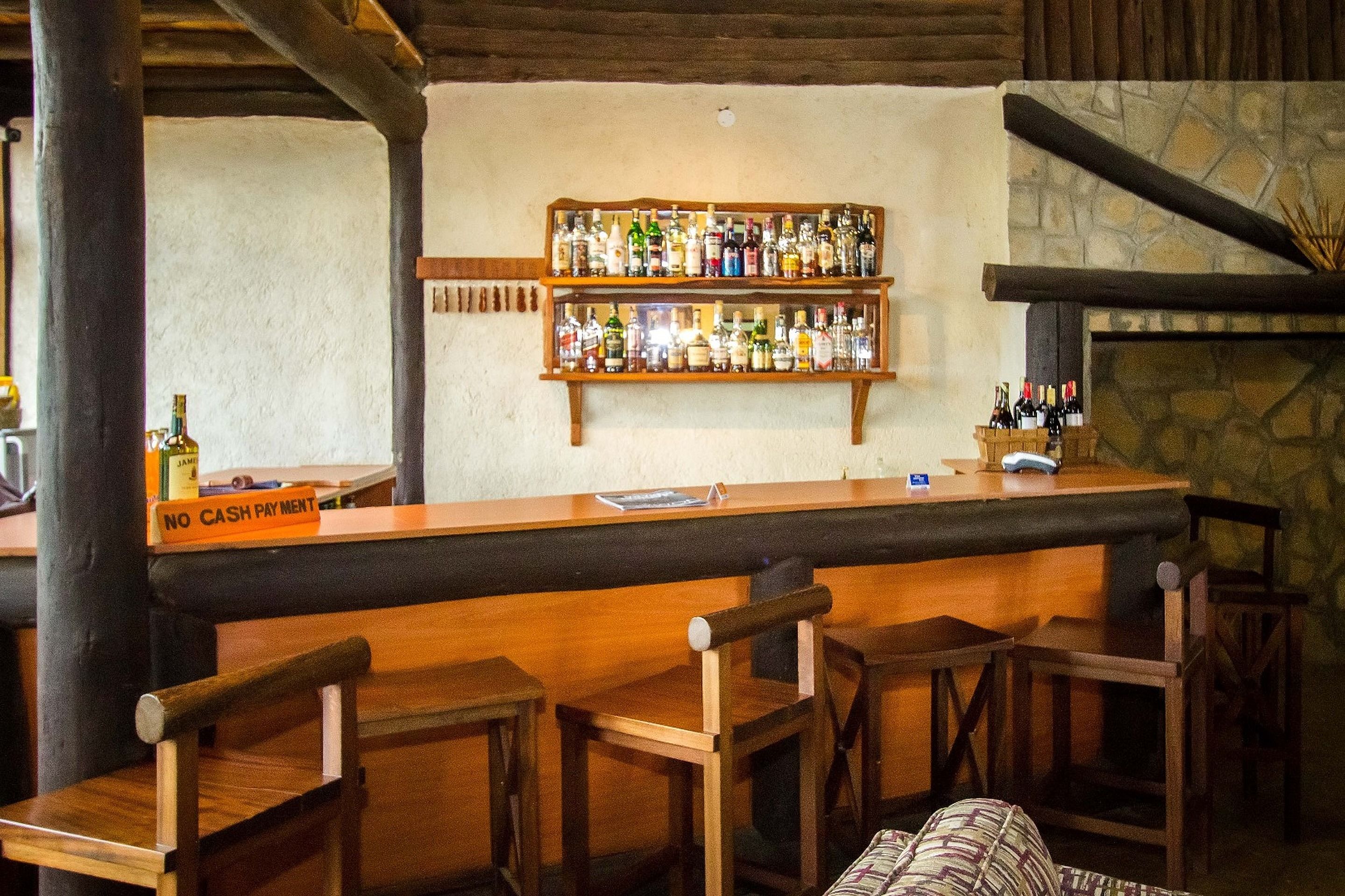 A bar area with wooden chairs and a 'NO CASH PAYMENT' sign, shelves with bottles, and a stone wall.