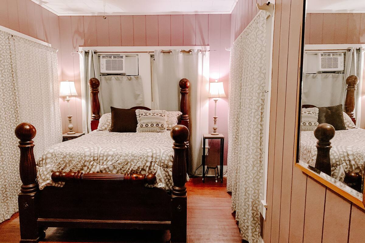 Un dormitorio con una cama de madera con dosel, dos lámparas de noche, una mesita con una silla y un espejo en la pared.
