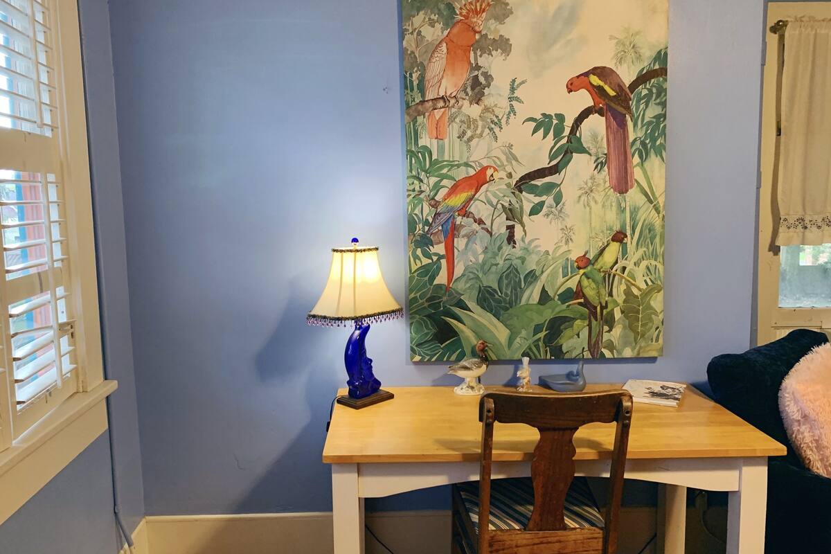 Una habitación con una mesa de madera, una silla, una lámpara y un cuadro de pájaros en la pared.