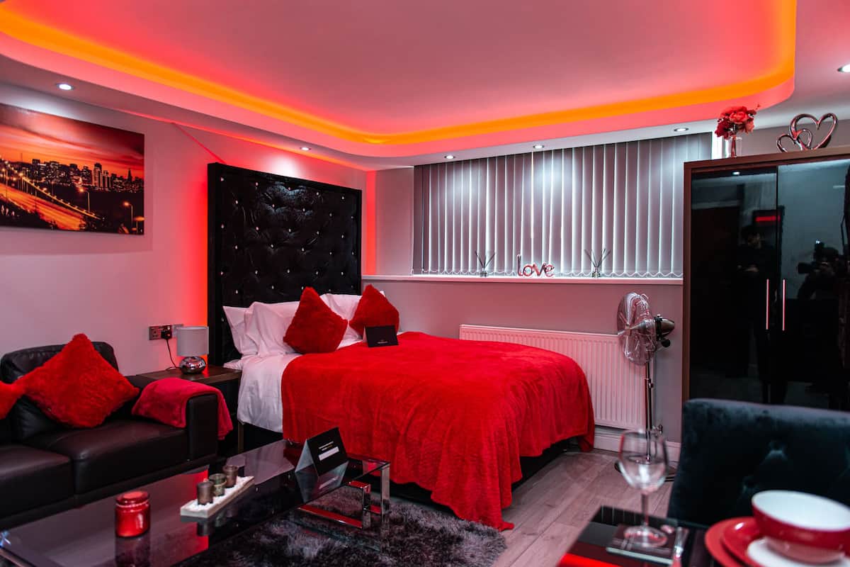 A modern bedroom with a red-themed bed, a black leather sofa, and a cityscape painting on the wall.
