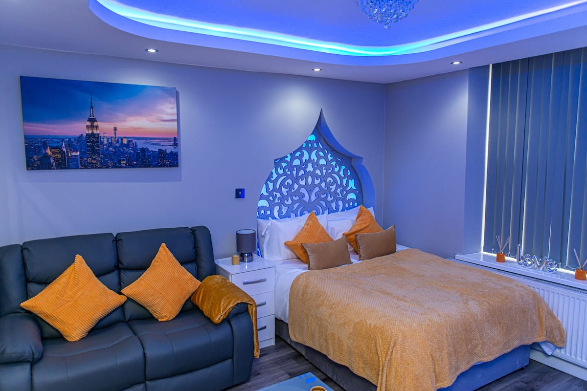 A modern bedroom with a cityscape painting, a patterned headboard, a bed with orange bedding, a dark blue sofa with orange cushions, and a nightstand with a lamp.