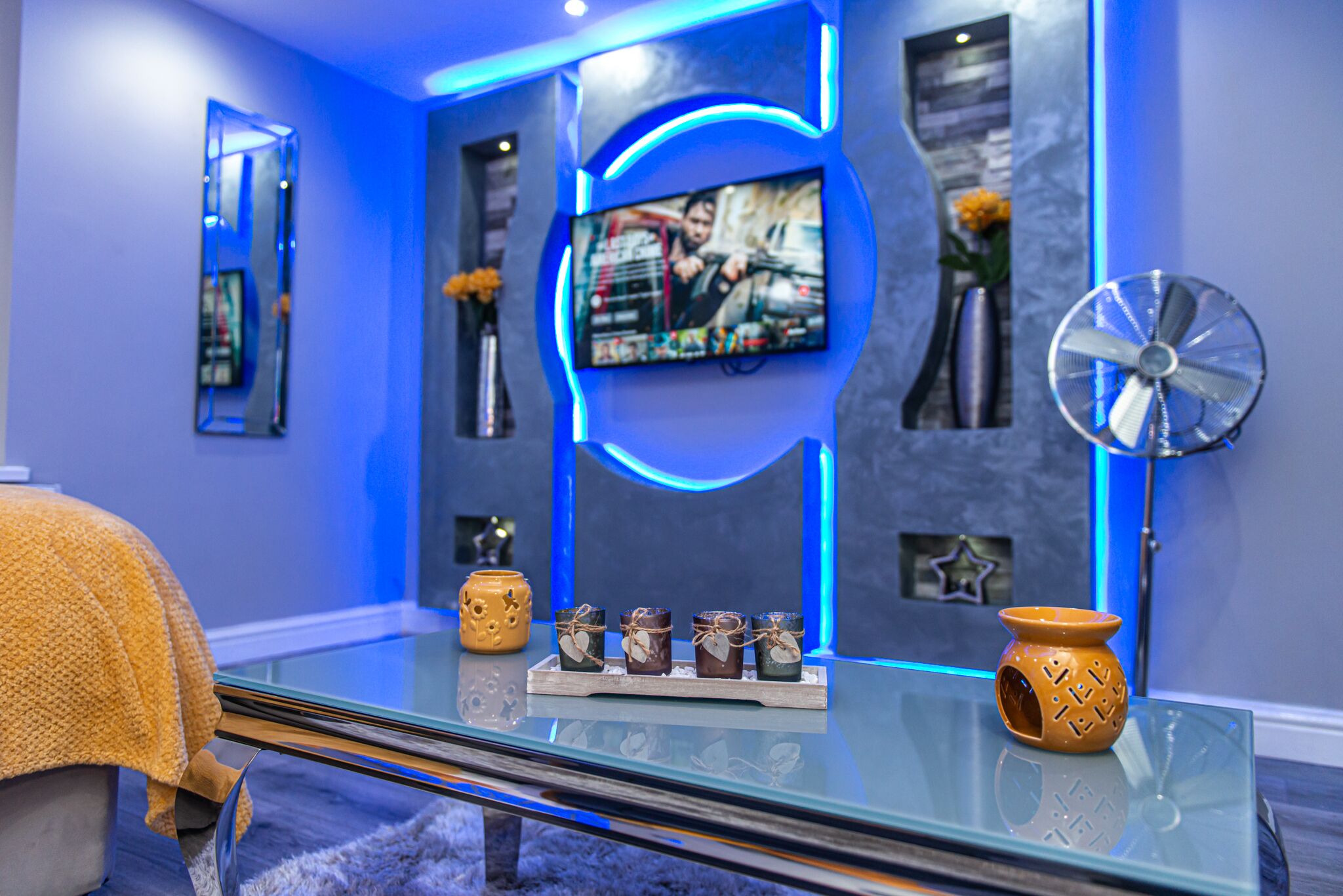 A modern living room with a glass coffee table, a flat-screen TV mounted on the wall, and decorative items including candles and a fan.