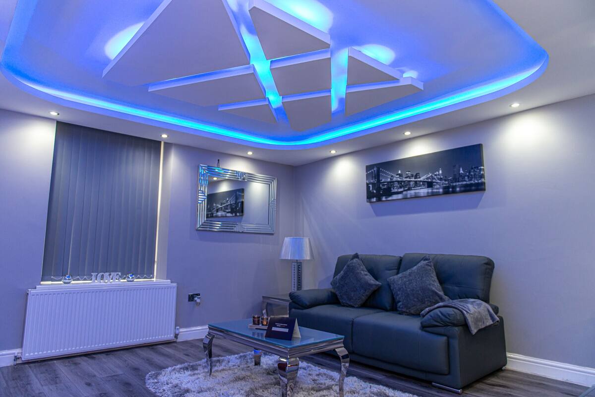 A modern living room with a blue-lit ceiling, a grey sofa, a glass coffee table, and a framed cityscape picture on the wall.