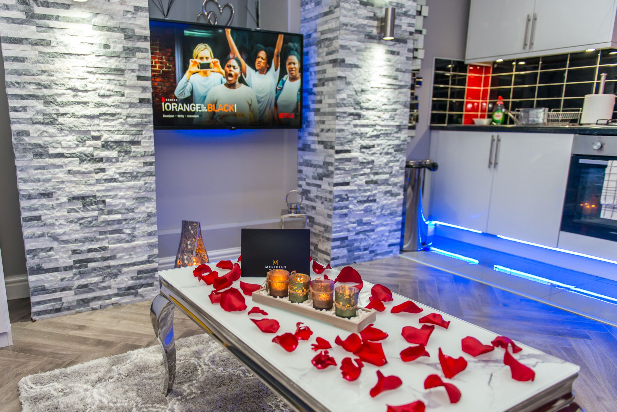 Una sala de estar moderna con una pared de piedra, un televisor de pantalla plana, una mesa de centro con velas y una cocina al fondo.