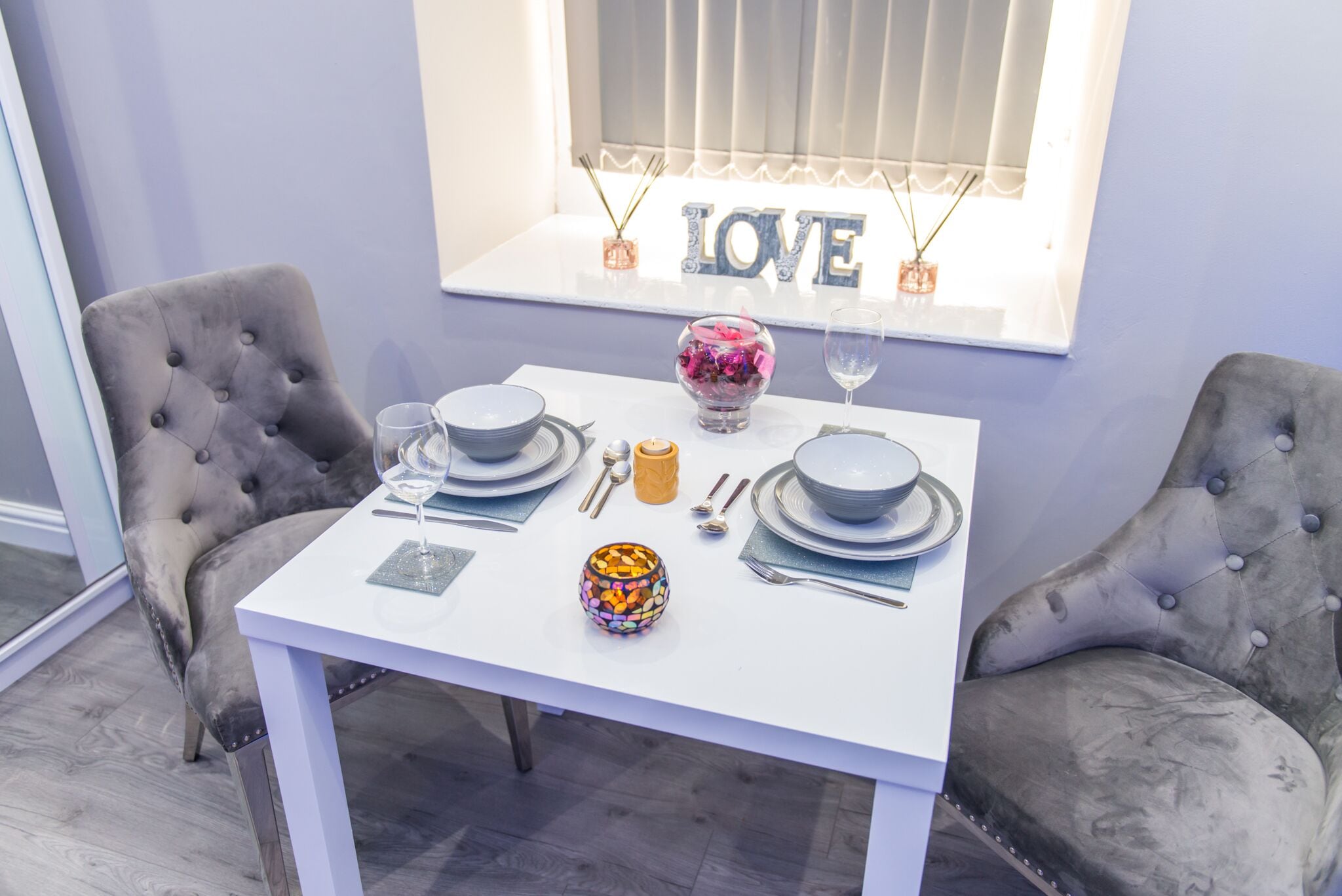 A dining table set with plates, cutlery, and a decorative bowl, flanked by tufted chairs, in a room with a window and a 'LOVE' sign.