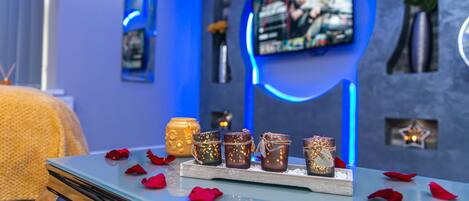 A spa area with a glass table displaying candles and rose petals, a television on the wall, and a blue ambient lighting.