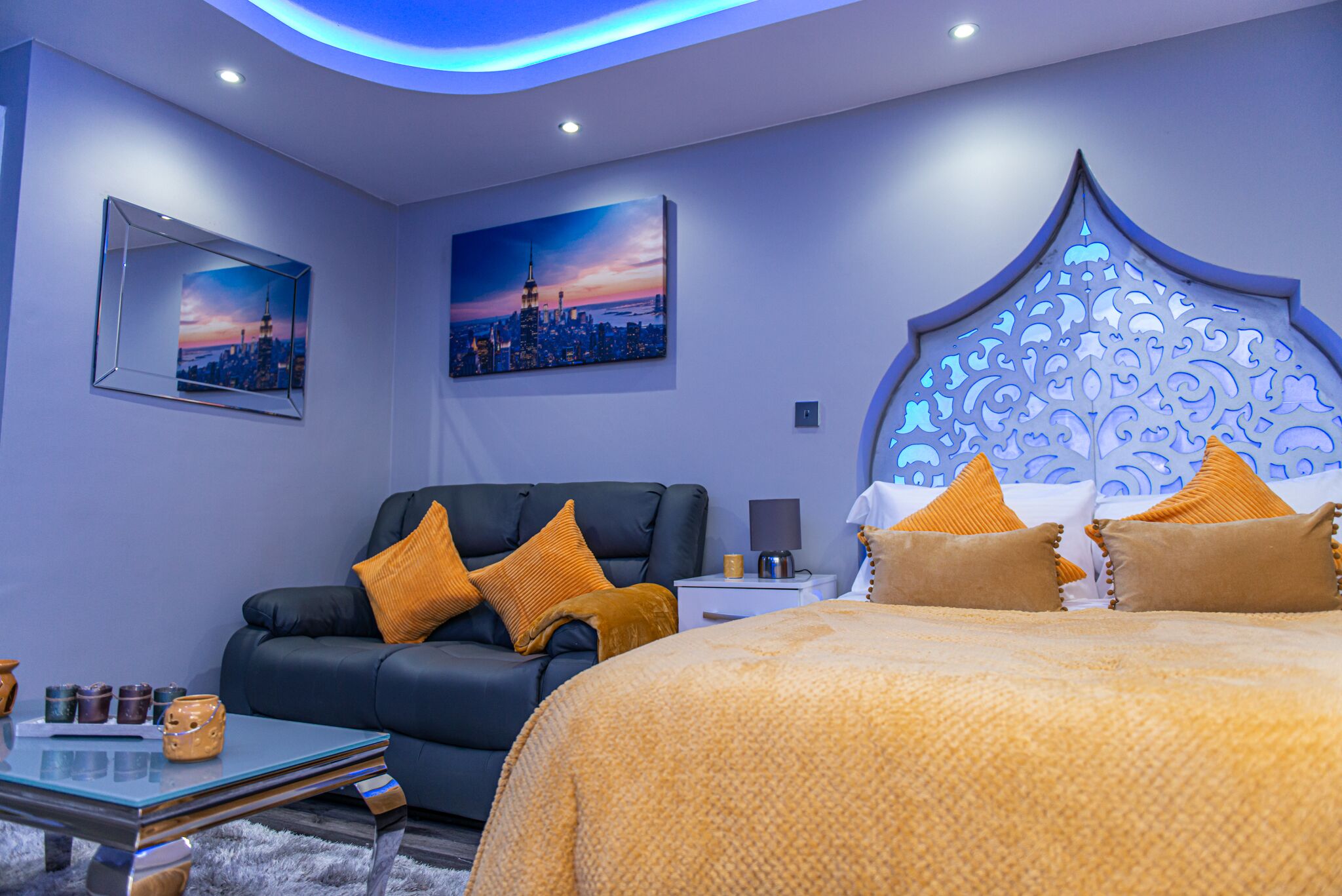 A modern bedroom with a cityscape painting, a black leather sofa, a bed with orange bedding, and a decorative headboard.