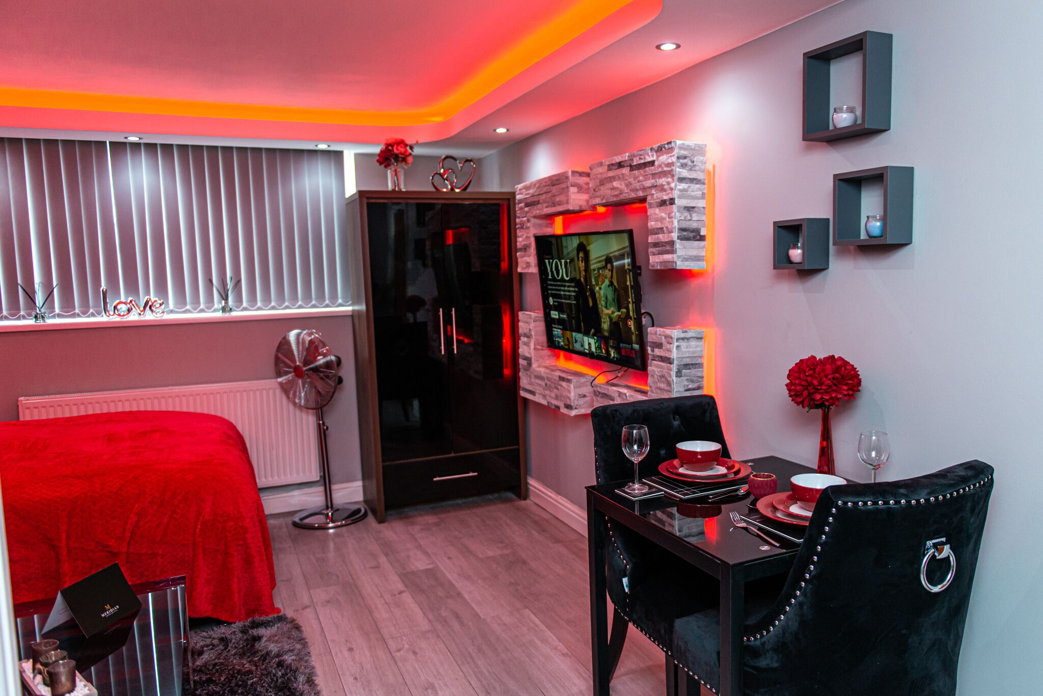 A modern bedroom with a red bedspread, a black wardrobe, a dining table set with red and white plates, and a wall-mounted TV.