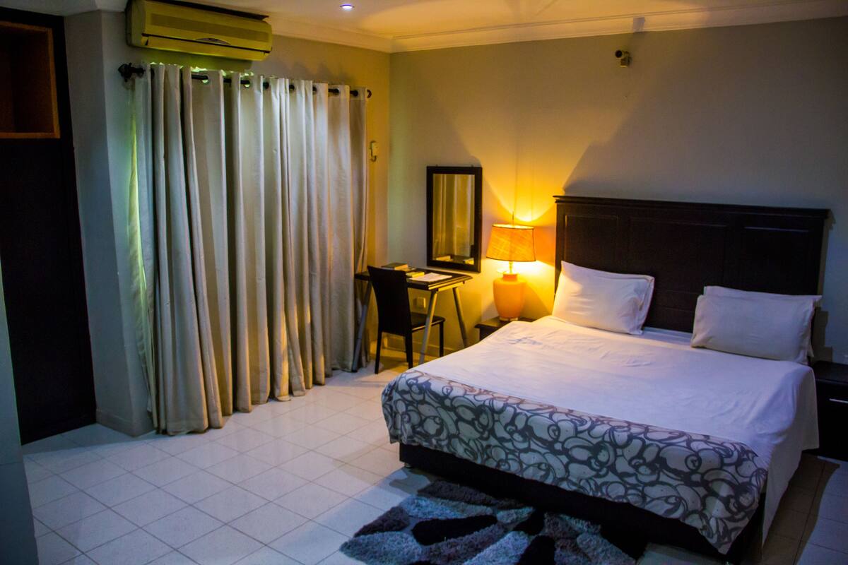 A hotel room with a bed, a desk, a chair, a lamp, and curtains.