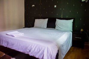 A neatly made bed with white linens and a folded towel, a dark headboard, and a nightstand with a lamp. - Lekki Heights Hotel (Lekki)
