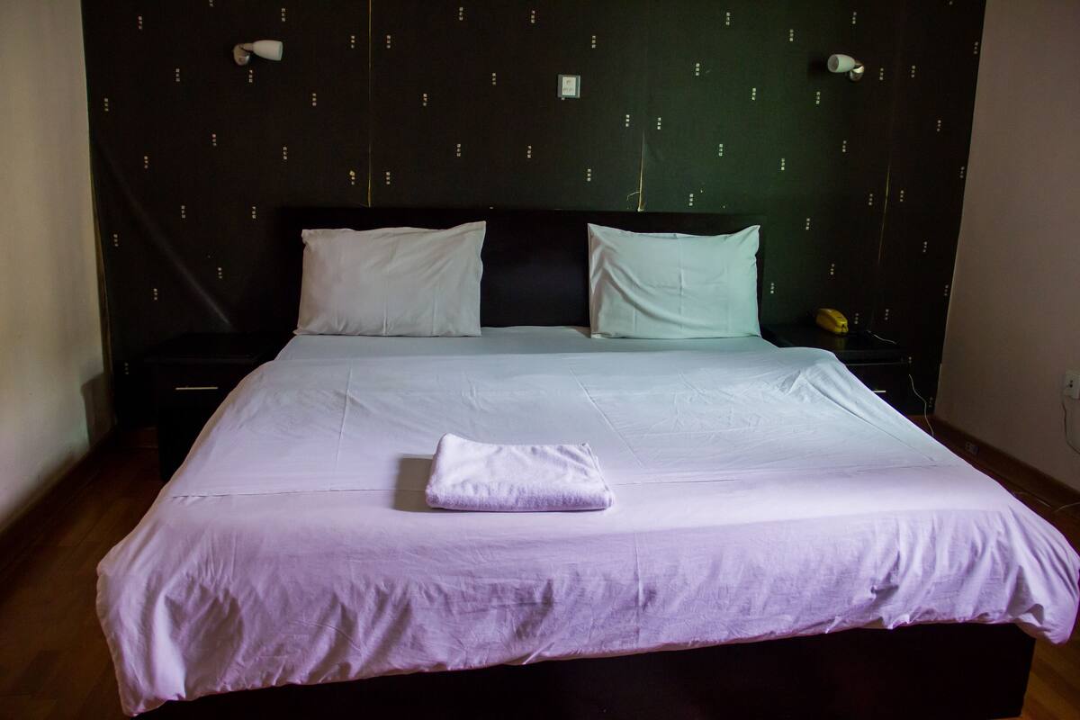 A neatly made bed with white linens and a folded towel, a black headboard, and a small bedside table with a lamp.