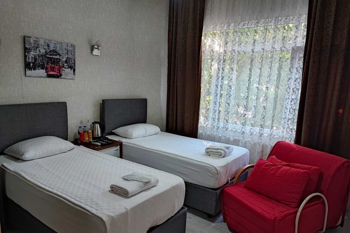 Ein Hotelzimmer mit zwei Einzelbetten, einem roten Sessel, einem kleinen Tisch mit Wasserkocher und einer Flasche Wasser sowie einem Fenster mit Vorhängen.