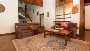 Ein geräumiges Wohnzimmer mit zwei braunen Sesseln, einem Couchtisch aus Holz und einem großen, gemusterten Teppich. Im Hintergrund ist eine Treppe zu einer oberen Ebene zu sehen.