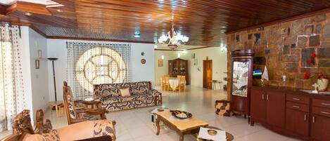 A spacious living room with a wooden ceiling, a patterned sofa, a dining table with chairs, and a stone wall with a cabinet.