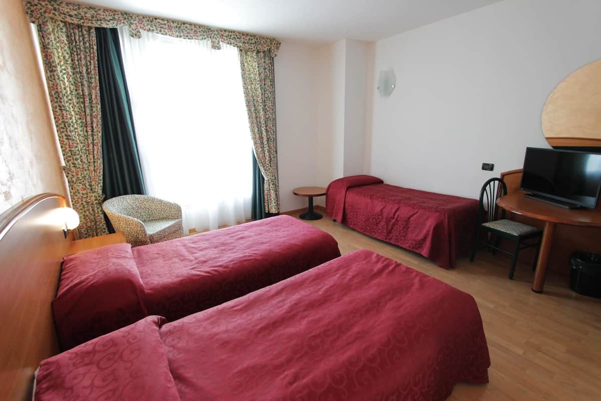 Ein Hotelzimmer mit zwei Einzelbetten, einem kleinen runden Tisch, einem Sessel, einem Fernseher und einem Fenster mit Vorhängen.
