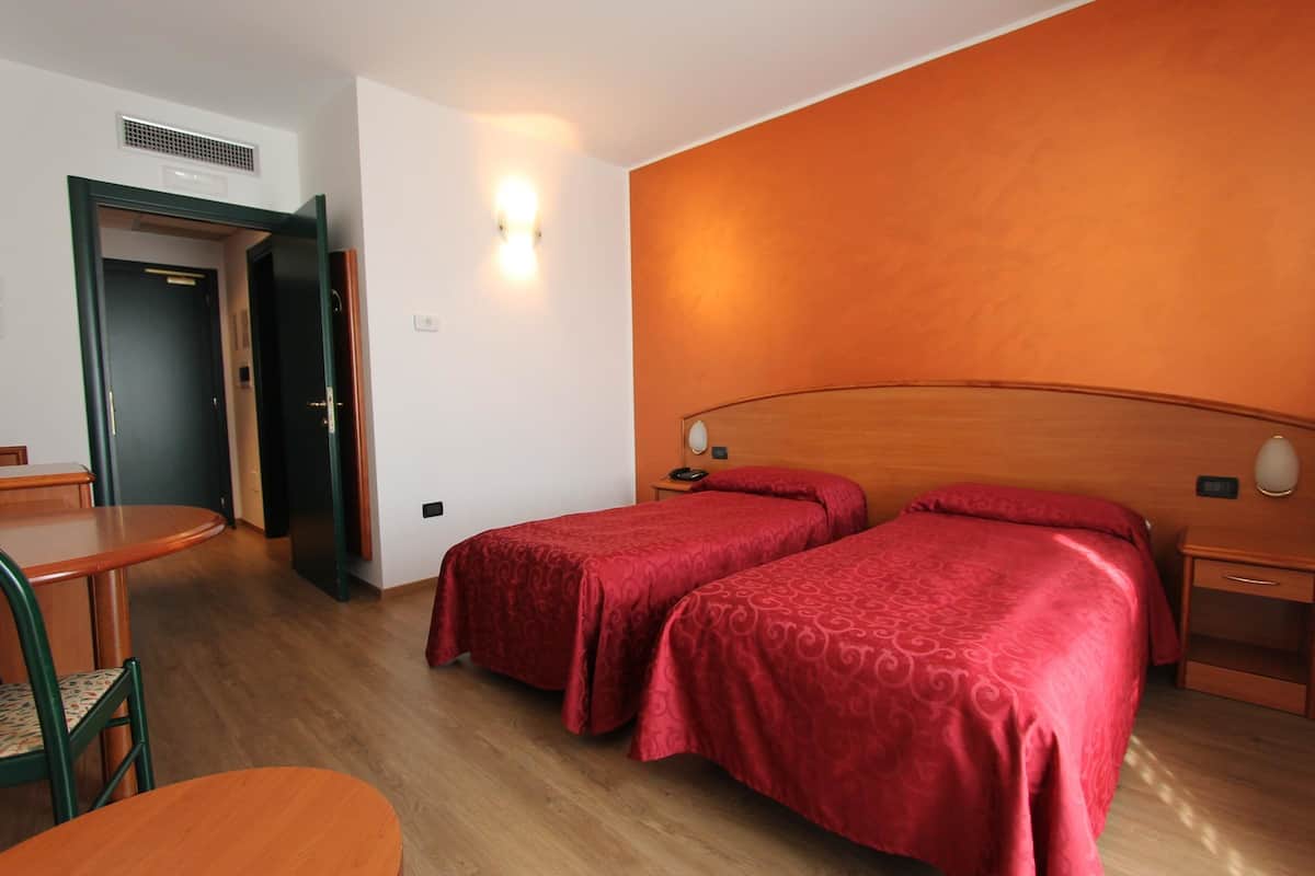 Ein Hotelzimmer mit zwei Einzelbetten, einem Holzkopfstück, einem Nachttisch mit Lampe, einem kleinen Tisch mit Stuhl und einer Tür zu einem weiteren Raum.